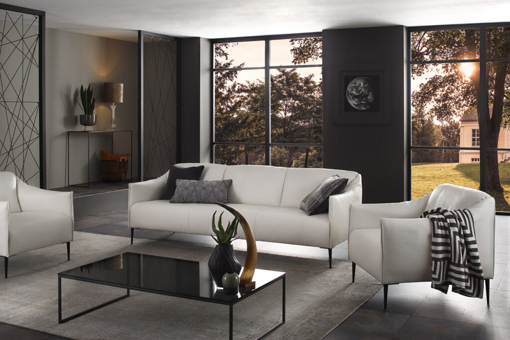 Sofa uns Sessel Modell 15350 in einer Raumszene bei Tageslicht, in weißem Leder, mit schwarzem Metallfuß, Schrägansicht, moderner Wohnstil, dezent dekoriert, offen und hell mit großen Fenstern und Vinylboden. Der Sessel ist seitlich an der Couch positioniert und fügt sich harmonisch ins Gesamtbild ein