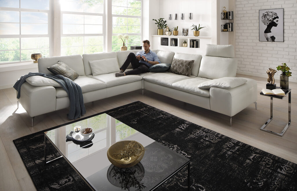 Eckkombination Modell 16540 in einer Raumszene bei Tageslicht in weißem Leder, mit chromglänzendem Metallfuß, Schrägansicht mit Flexibler Steckkopfstütze, moderner Wohnstil, dezent dekoriert, offen und hell mit großen Fenstern und holzoptik Vinylboden
