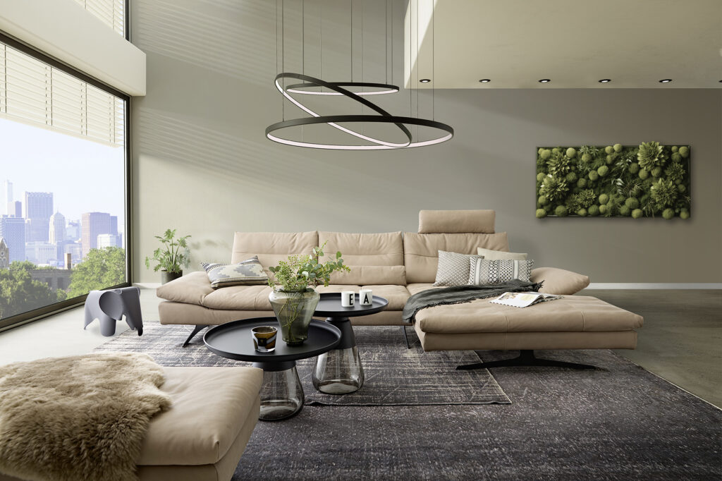 Sofa-Longchair-Kombination Modell 16741 in einer Raumszene bei Tageslicht in beigem Leder, mit schwarzer Metallkufe, Kopfstütze, Frontansicht mit Seitenteilverstellung, Flexibler Steckkopfstütze und Hocker im Vordergrund im Anschnitt. Nierenkissen am Rücken platziert, Moderner Eigenheim-Stil, großzügig, edel, gehoben und minimalistisch. Mit Holz-Akustikwand im Hintergrund und markanter Deckenleuchte in Metallopik, anthrazitfarbene große Teppiche, bodentiefes großes Fenster